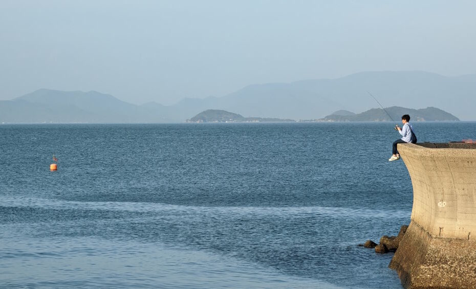 Young man fishing on an anti-tsunami wall on the shore of the Seto Inland Sea. We can see the islands of Oshima and Shodoshima in the background.