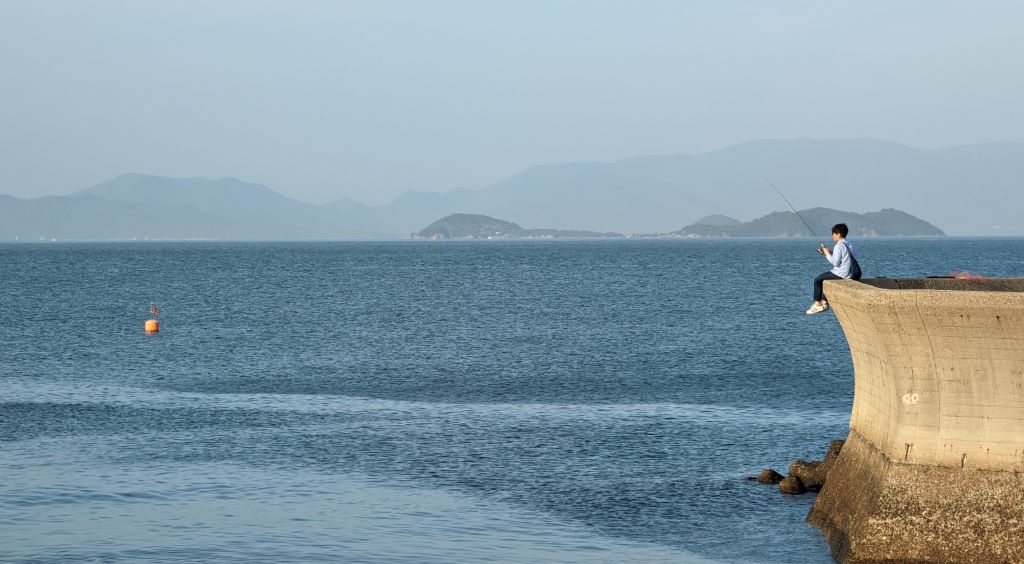 Young man fishing on an anti-tsunami wall on the shore of the Seto Inland Sea. We can see the islands of Oshima and Shodoshima in the background.